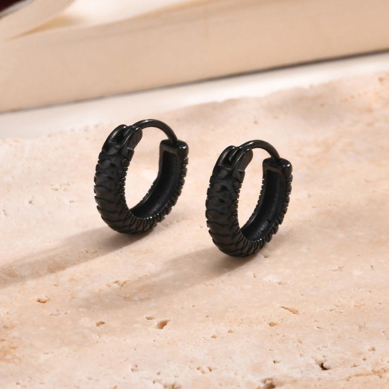 Paire de boucles d'oreilles anneaux noires en acier inoxydable posée sur un fond clair minimaliste, mettant en valeur leur finition texturée et élégante.