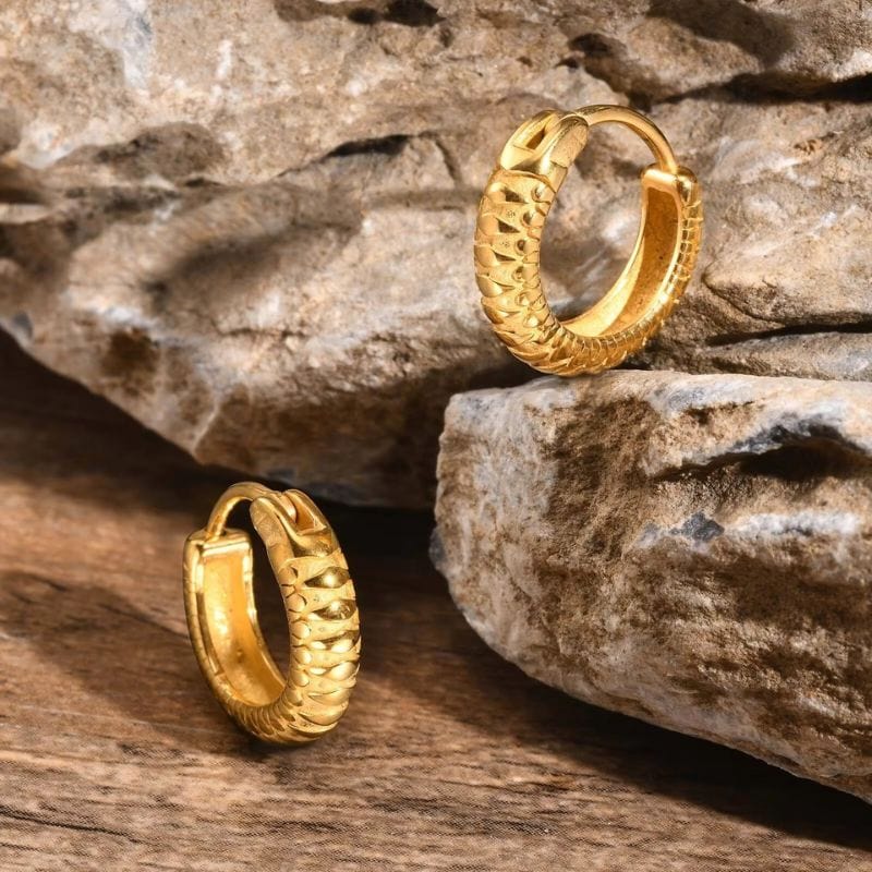 Gros plan sur paire de boucles d'oreilles anneaux en acier inoxydable doré avec motifs en relief posées sur un décor naturel en pierre