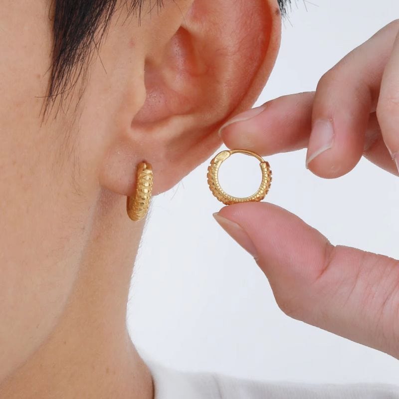 Boucles d'oreilles anneaux dorés en acier inoxydable pour homme, avec motif texturé, portées à l'oreille et présentées dans la main.