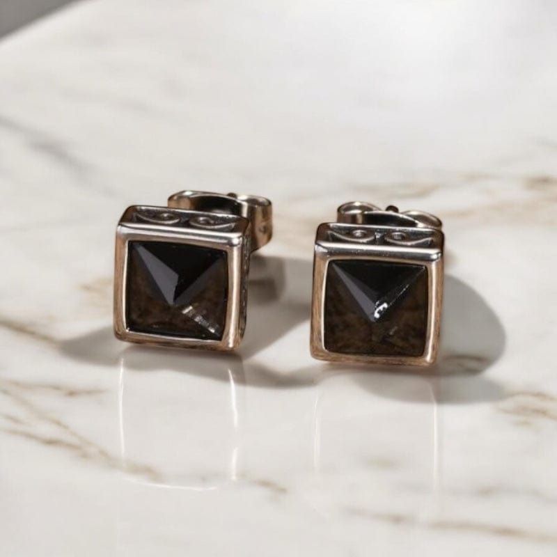 Paire de boucles d’oreilles clou en acier avec pierre noire carrée, exposées sur une surface en bois.