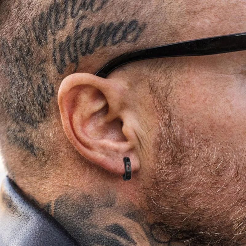 Boucle d’oreille pour homme en forme d’anneau noir avec croix – portée sur oreille tatouée.