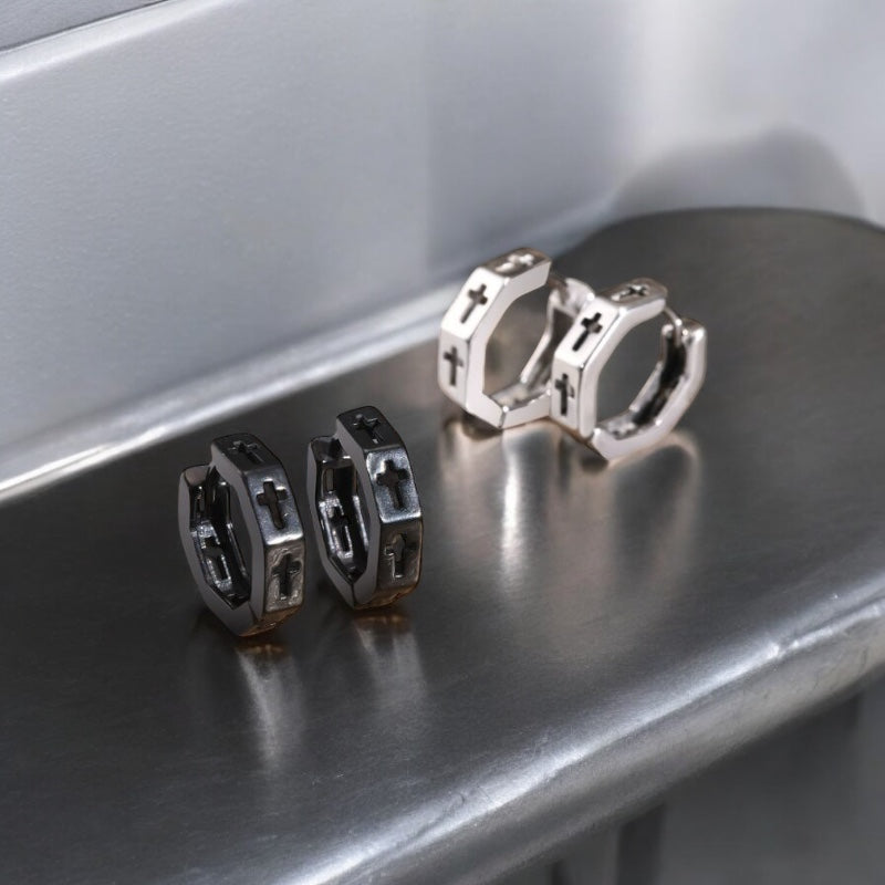 Paire de boucles d’oreilles créoles homme en acier inoxydable noir et argent avec croix gravée, posées sur fond en bois 
