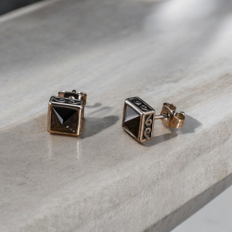 Paire de boucles d’oreilles carrées pour homme en acier inoxydable avec pierre noire pyramidale, posées sur surface boisée. 