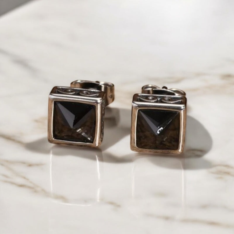 Paire de boucles d’oreilles clou en acier avec pierre noire carrée, exposées sur une surface en bois. 
