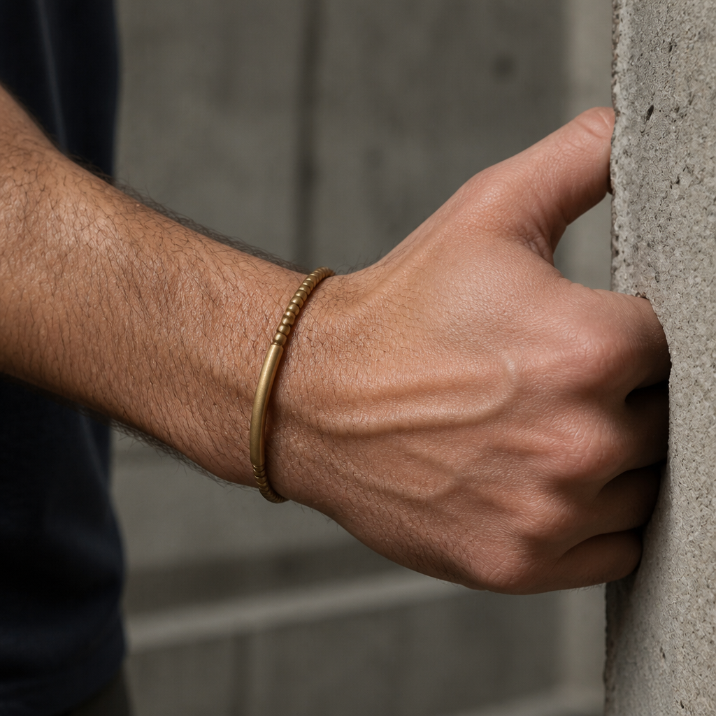 Bracelet homme doré au poignet, main posée sur un mur de béton