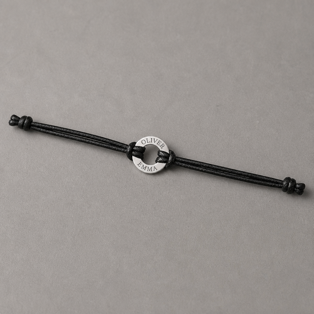 Bracelet homme à cordon noir posé à plat, anneau d'acier gravé de deux prénoms et nœuds coulissants visibles